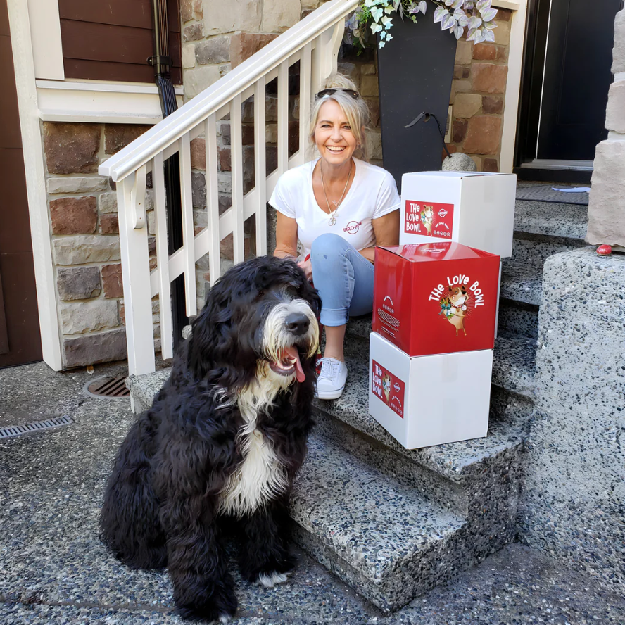 Smiling woman with her large black-and-white dog sitting on front steps beside stacked boxes of The Love Bowl—nutritious, pet-friendly meals made with wholesome, sustainable Maia Farms ingredients.