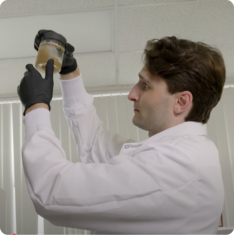 Maia Farms scientist in a white lab coat and black gloves inspects a jar of mycelium culture under fluorescent light, representing Maia Farms’ precision fermentation research in action.
